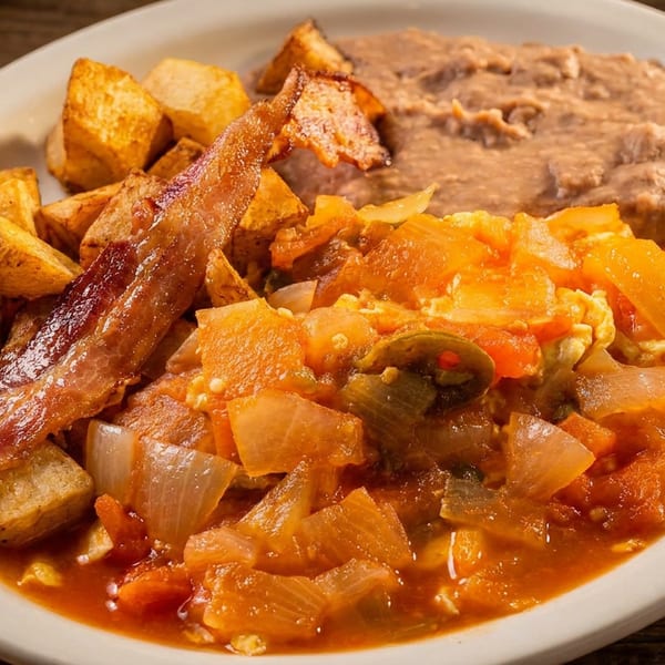 Plate of scrambled eggs with tomatoes, onions, peppers, bacon, roasted potatoes, and refried beans on a wooden table.