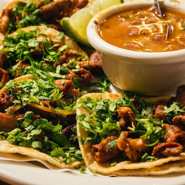 Three meat tacos topped with cilantro and onions, served with lime and a bowl of reddish salsa on a white plate.