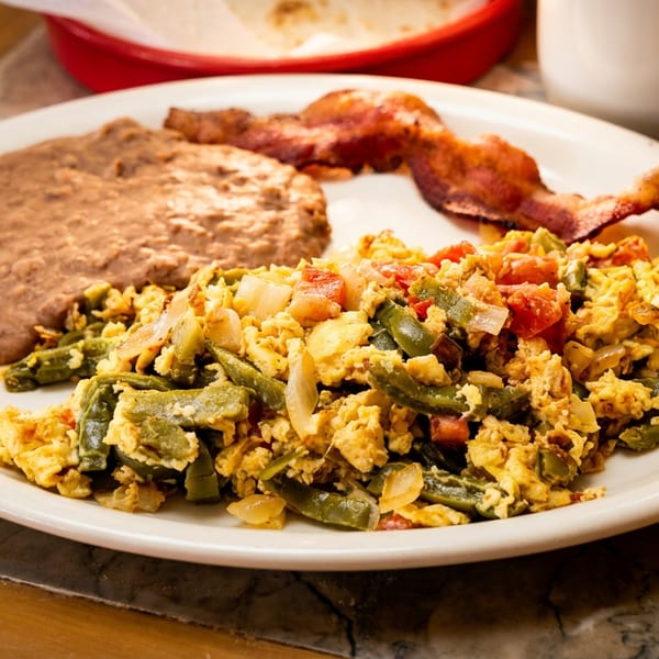Plate of scrambled eggs with peppers, onions, refried beans, and bacon on a restaurant table.