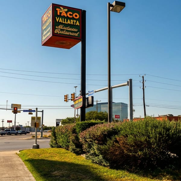 Taco Vallarta Mexican restaurant sign overlooking a sunny urban intersection with traffic lights, cars, shrubs, and clear blue sky.